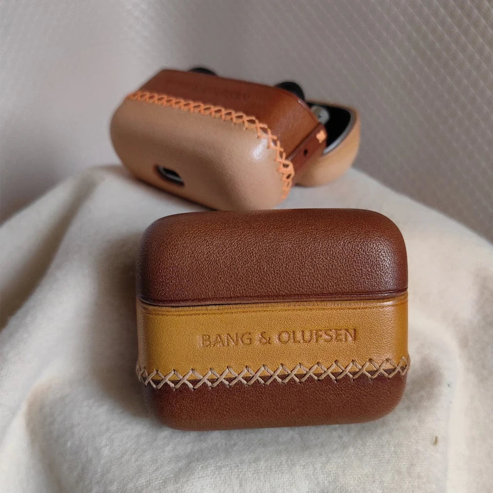 Detailed view of handmade dual-tone brown and beige leather case for Bang &amp; Olufsen Beo Grace earbuds charging case, showing the embossed logo and contrasting cross-stitch detail on a textured fabric background.