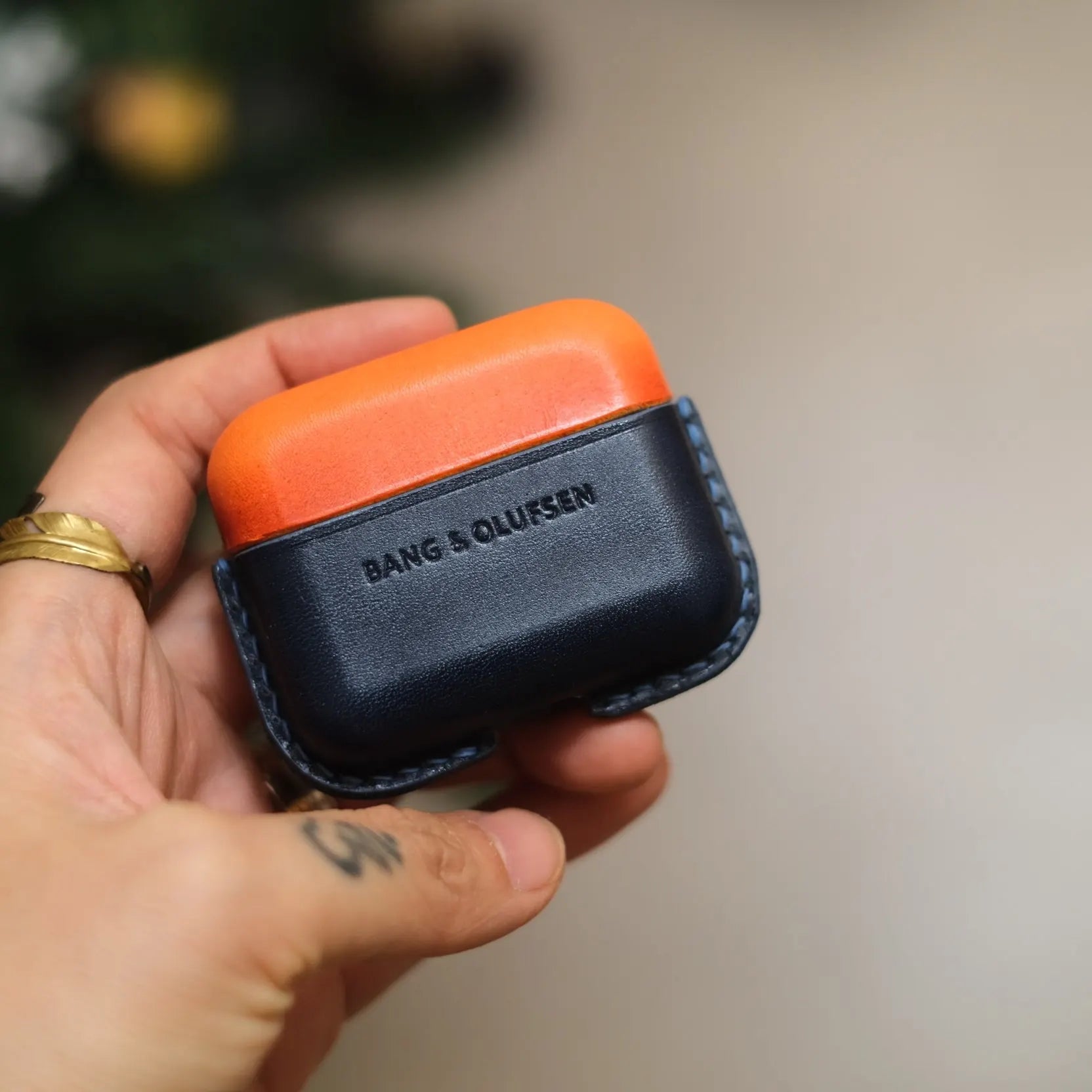 Hand holding an orange and navy blue two-tone handcrafted leather protective case for Bang & Olufsen Beo Grace, featuring debossed BANG & OLUFSEN logo.