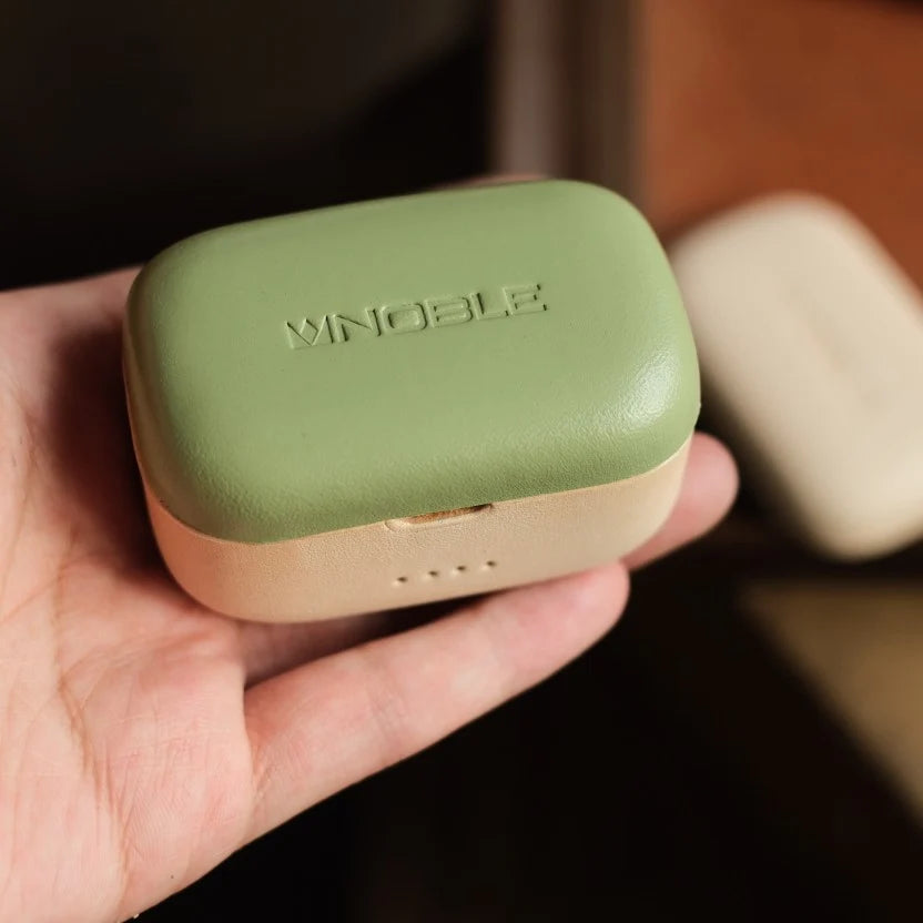 Eine Hand hält eine personalisierte Schutzhülle aus echtem Leder für Noble FoKus Prestige in Avocadogrün und Beige mit geprägtem Schriftzug 'NOBLE' auf der Oberseite.
