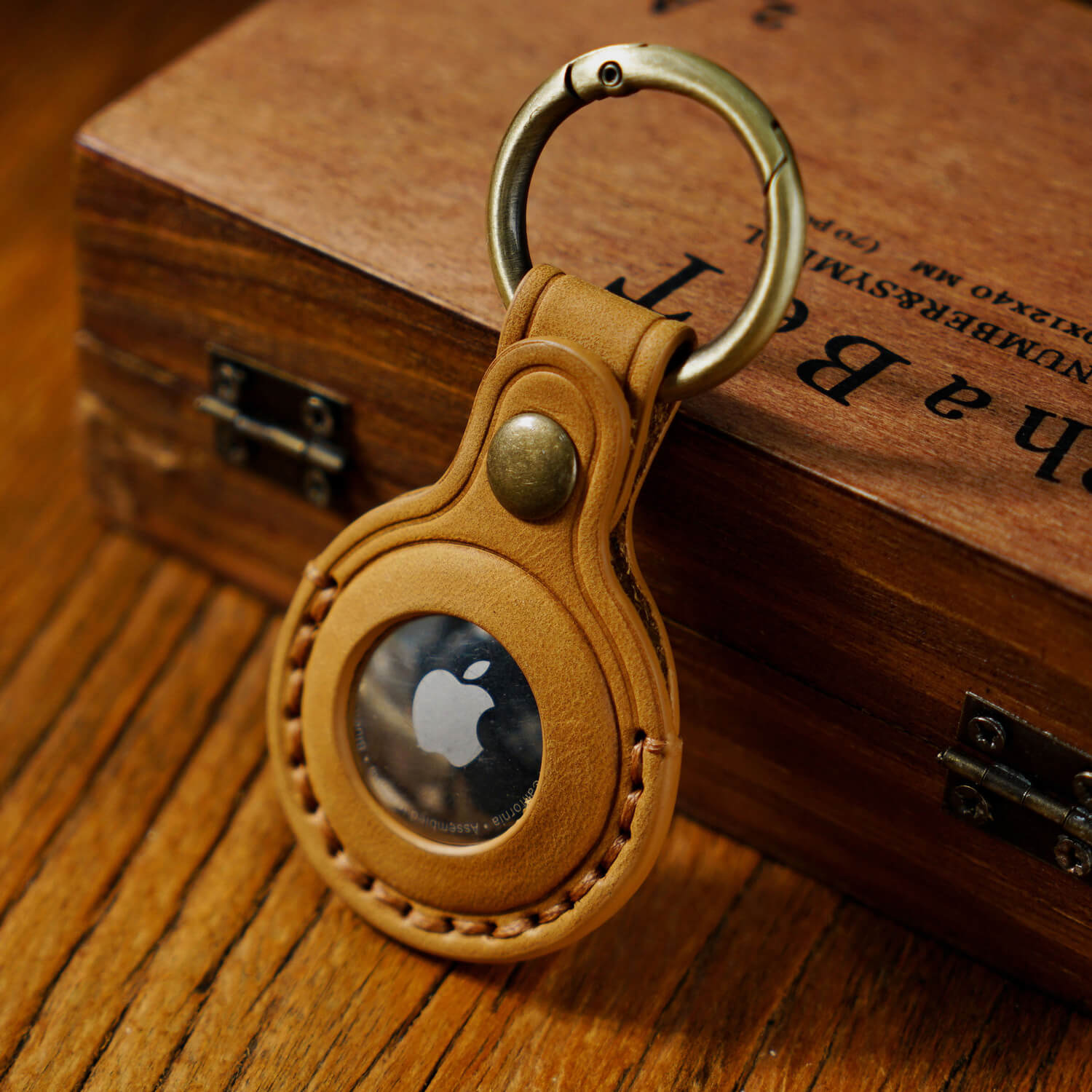 A stitched leather case holding an Apple AirTag is placed against a wooden box; The leather case has a vintage brass ring.