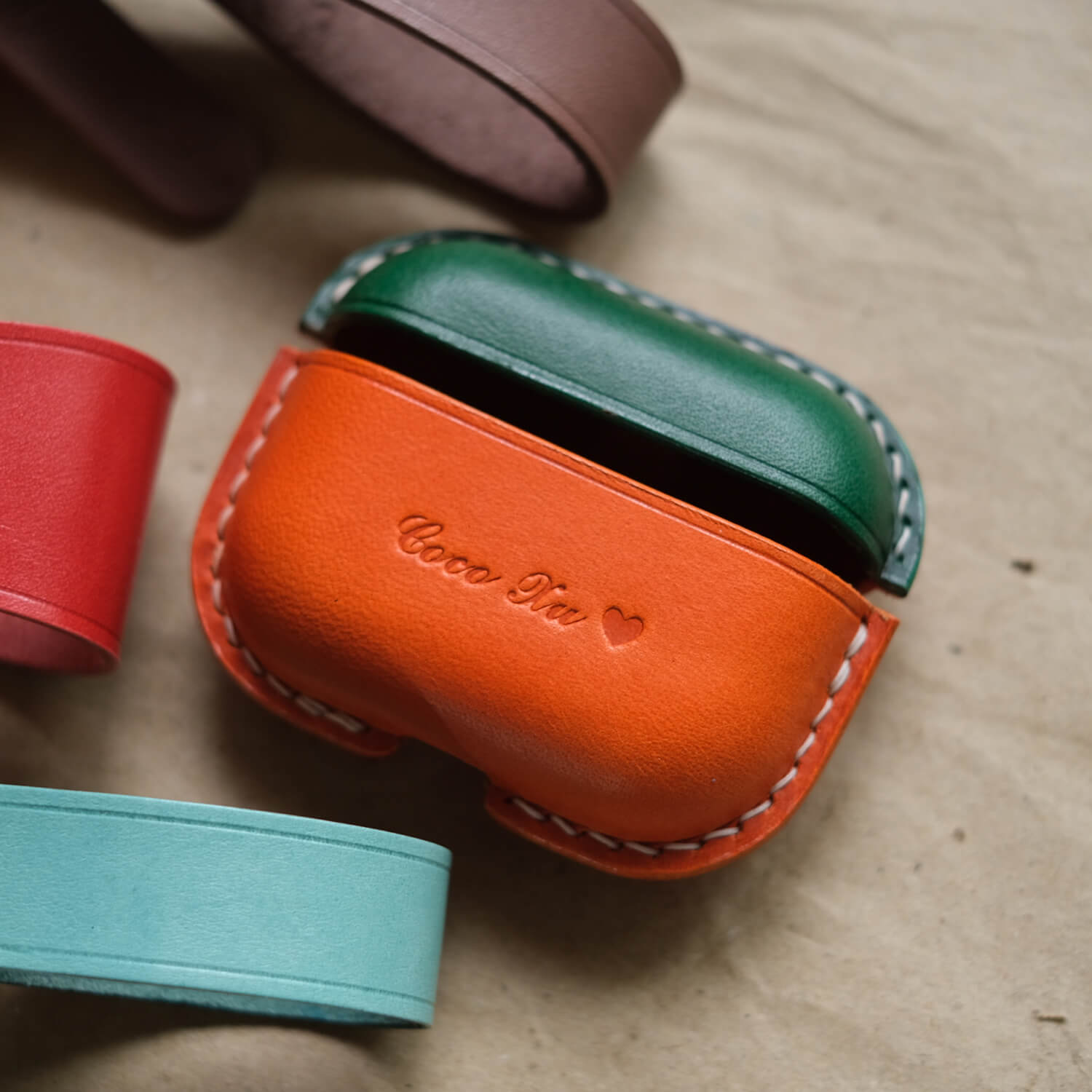 Two-tone handcrafted leather earbuds case for AirPods Pro 3 in green and orange with custom engraved text, showing premium stitching and personalized design options