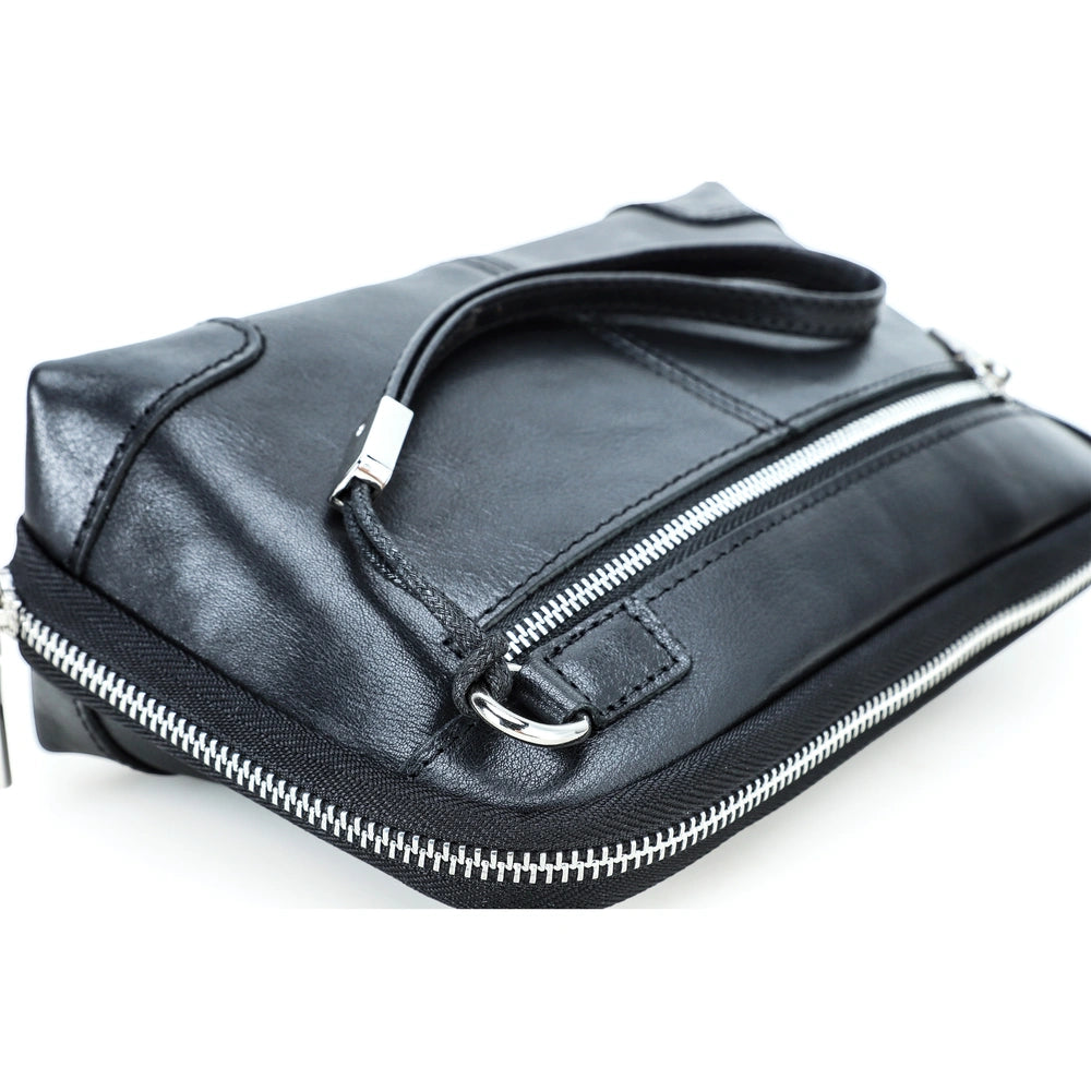 Close-up angle view of a handmade black vegetable-tanned leather clutch bag, highlighting the silver zipper closures, front pocket with metal D-ring, and wristlet strap.