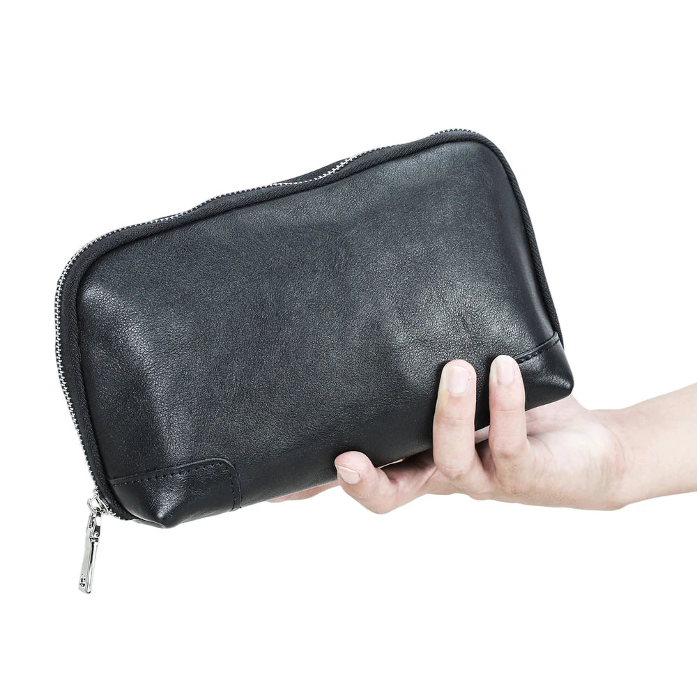 A hand holding a black handmade vegetable-tanned leather clutch pochette bag from the bottom, demonstrating its size, supple texture, and zipper closure.