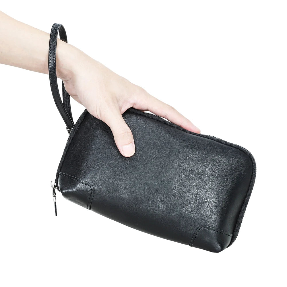 A hand holding a black handmade vegetable-tanned leather clutch bag with the wristlet strap worn around the wrist.