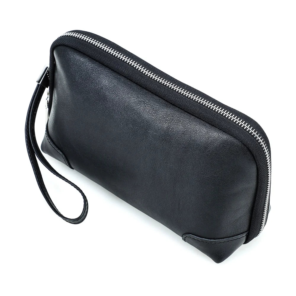 Side and back angle view of a handmade black vegetable-tanned leather clutch bag, highlighting the structured profile, zipper closure, and wristlet strap attachment.