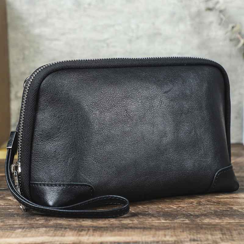 Handmade black vegetable-tanned leather clutch bag with zipper closure and wristlet strap resting on a rustic wooden surface against a concrete wall background.