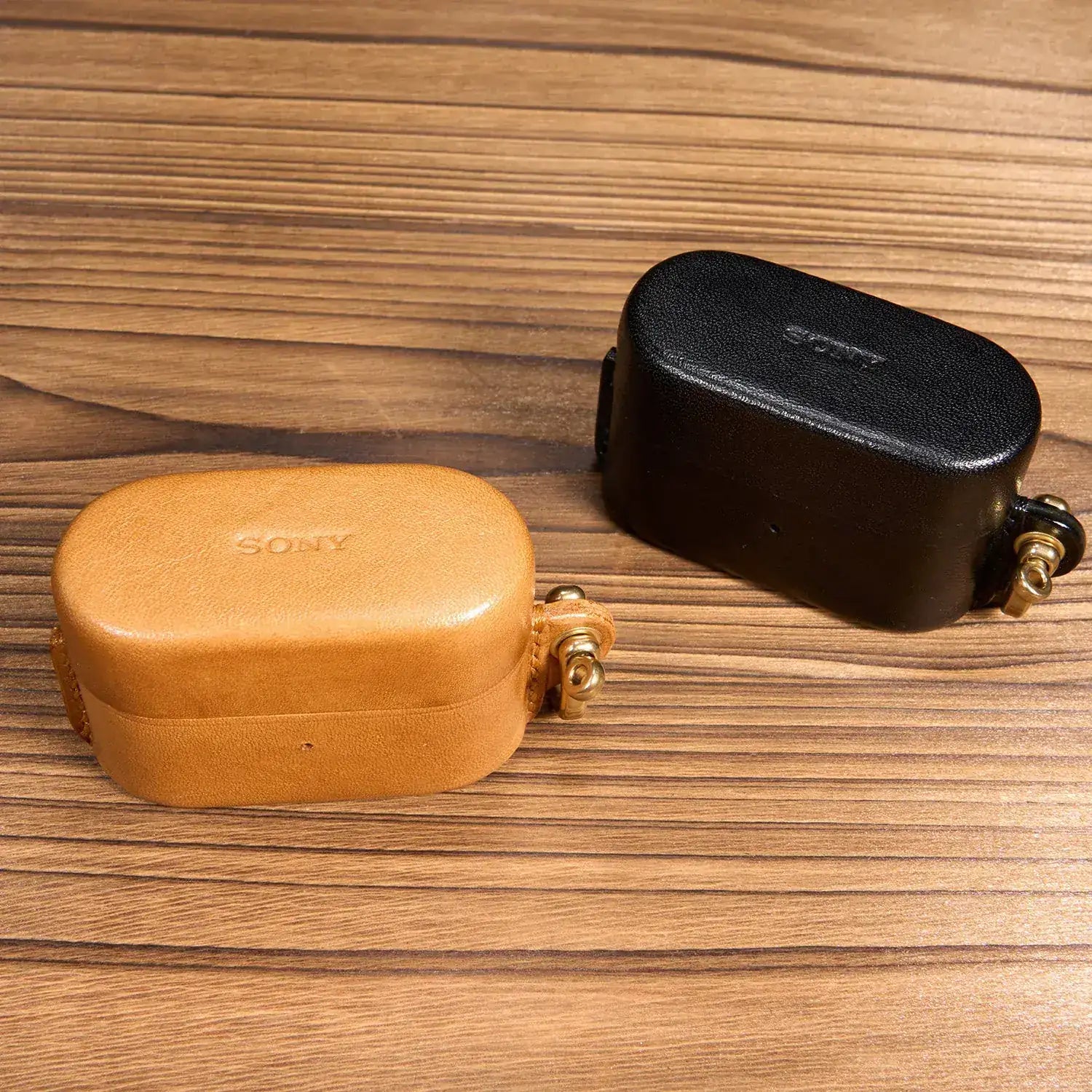 Confronto tra le custodie artigianali in pelle marrone e nera per Sony WF-1000XM6 su superficie in legno, che mostra le trame pregiate.